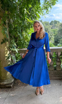 Katya wearing a blue dress, standing infront of a leafy backdrop