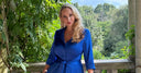 Katya wearing a blue dress, standing infront of a leafy backdrop