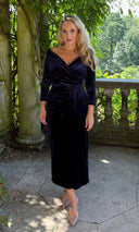 Katya wearing a  navy velvet Claridges wrap dress, standing on a stone balcony surrounded by lush green foliage.