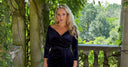 Katya wearing a  navy velvet Claridges wrap dress, standing on a stone balcony surrounded by lush green foliage.
